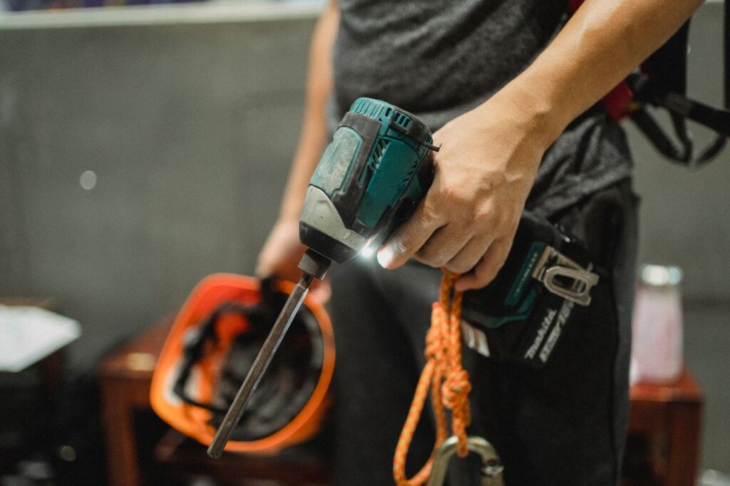 Crop unrecognizable mechanic wearing safety belt standing with switched on professional screwdriver and orange helmet before working indoors